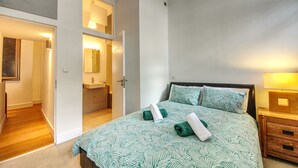 A bedroom with a bed, a nightstand, a bedside table, a mirror, and a bathroom visible through an open door.