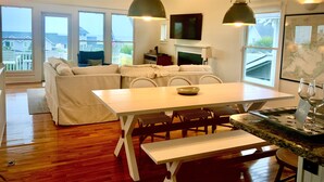 Ein modernes Wohnzimmer mit weißem Sofa, Esstisch und Stühlen. Es gibt einen Kamin, einen Fernseher und große Fenster mit Blick ins Freie.