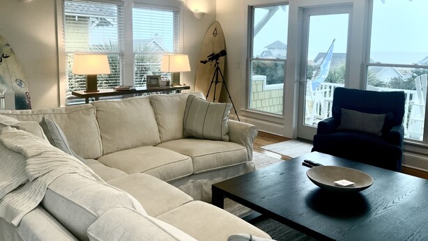 Ein Wohnzimmer mit einem beigefarbenen Ecksofa, einem dunklen Couchtisch aus Holz und einem blauen Sessel. Zwei Fenster mit Jalousien, ein Surfbrett und ein Stativ mit einer Kamera sind ebenfalls zu sehen.