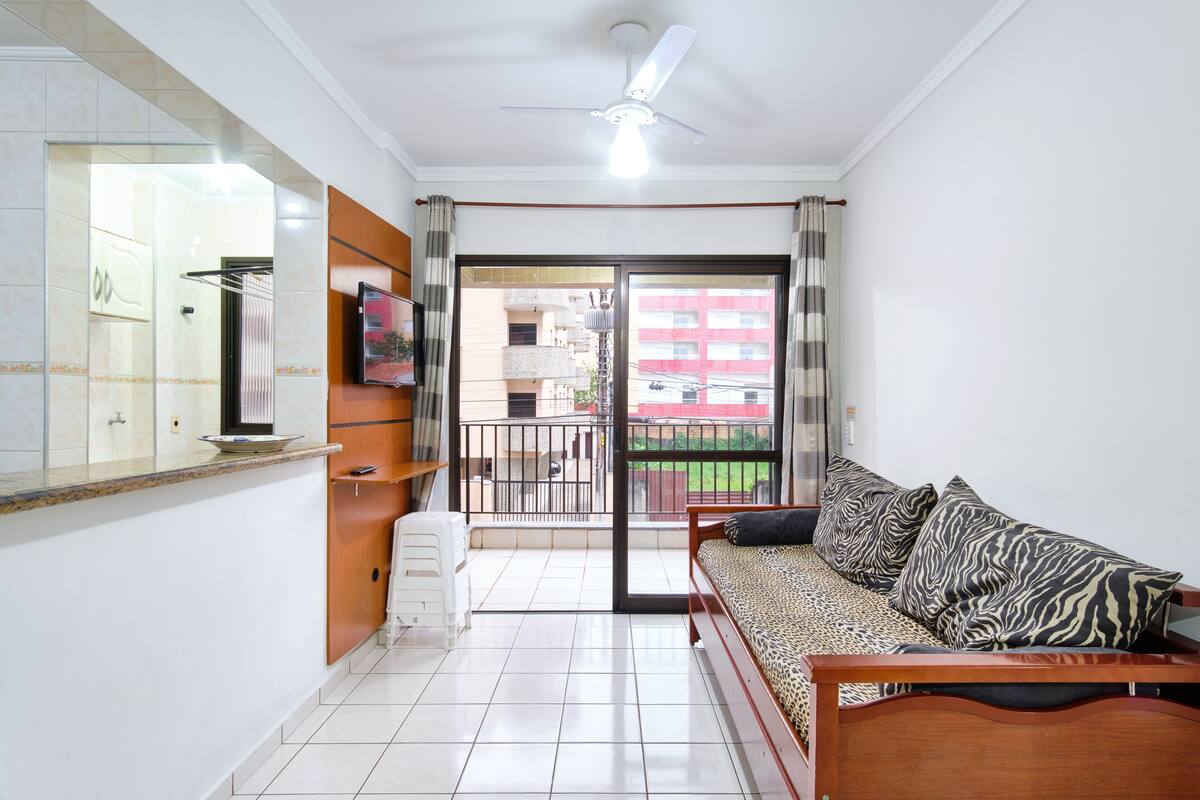 Una habitación con sofá, televisión, ventilador de techo, balcón y vistas a edificios, además de piso de baldosas.