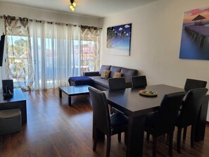 A living room with a sofa, coffee table, and dining table with chairs. There is a painting on the wall and a window with curtains.