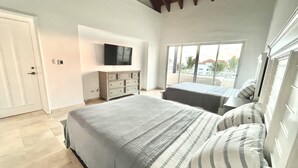 Ein Schlafzimmer mit einem großen Bett, einem an der Wand befestigten Fernseher und einem Fenster mit Blick auf Palmen.