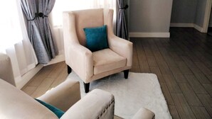 Ein Zimmer mit einem beigen Sessel, einem blauen Kissen, einem weißen Teppich und Holzdielen.