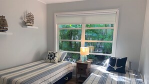 A bedroom with two beds, a nightstand with a lamp, and a window with a view of greenery. - Flip Flop 2 (Sanibel)