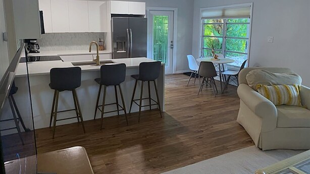 A modern kitchen with white cabinets, a stainless steel refrigerator, and a bar area with stools. There is a dining table with chairs and a sofa in the living area. - Flip Flop 2 (Sanibel)