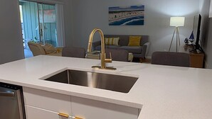 A modern kitchen with a stainless steel sink, white cabinetry, and a grey sofa in the background. - Flip Flop 2 (Sanibel)