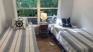 A room with two beds, a nightstand with a lamp, a window with a view of greenery, and a small wooden table. - Flip Flop 2 (Sanibel)