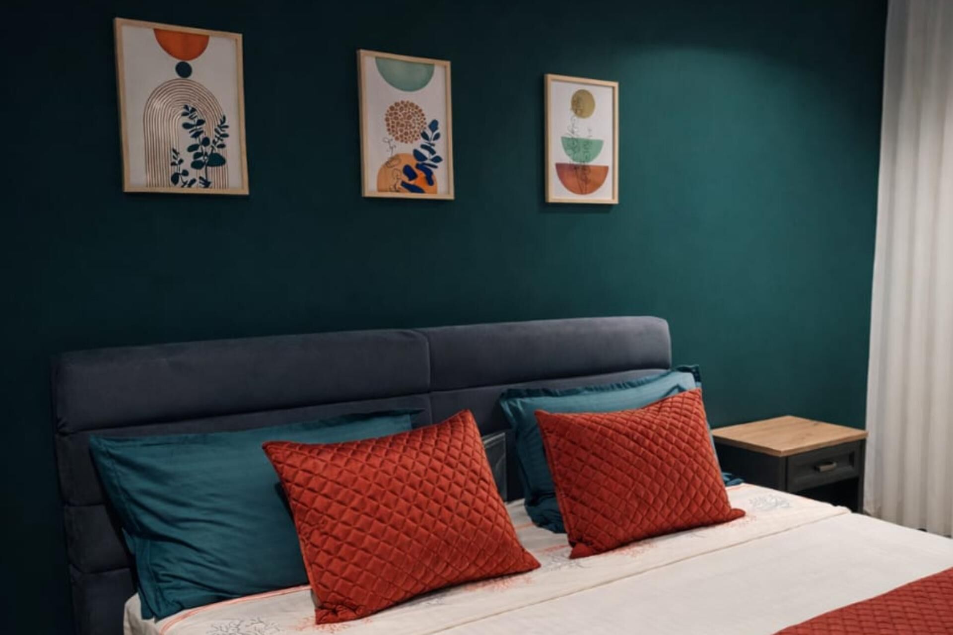 A bedroom with a dark headboard, teal and red pillows, and three framed artworks on the wall.