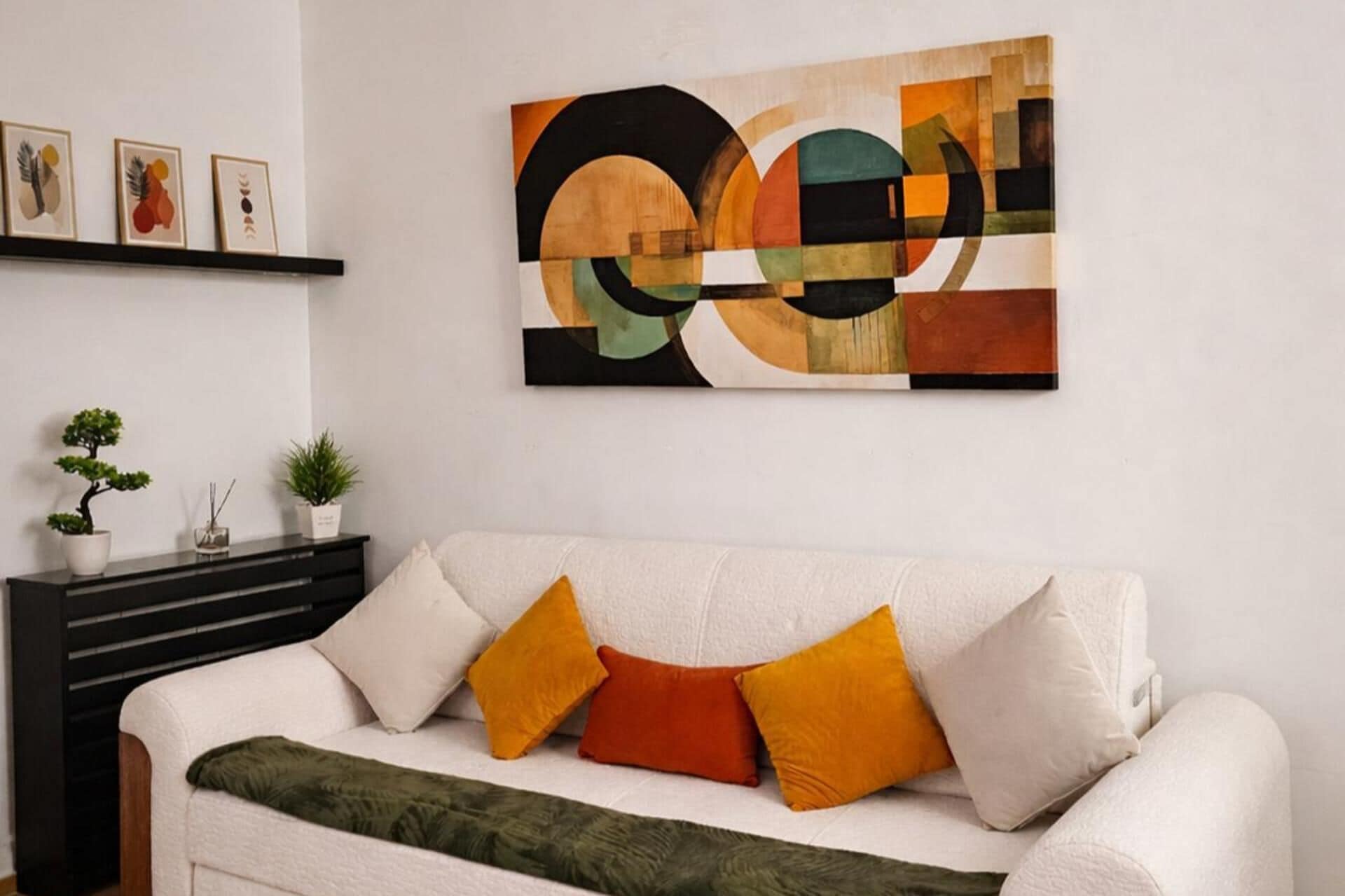Ein Wohnzimmer mit einem weißen Sofa, einem geometrischen Wandbild und einem kleinen Topf mit Pflanze auf einem schwarzen Konsolentisch.