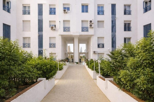 Un immeuble à plusieurs étages doté d’une entrée centrale, de climatiseurs et d’un chemin pavé bordé de verdure.