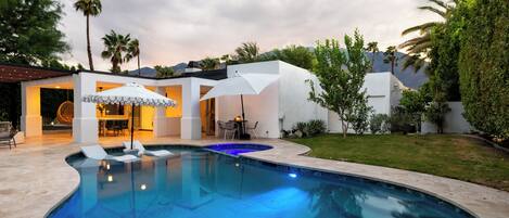 Piscina moderna ao ar livre com design curvo, rodeada por um terraço com assentos e guarda-sóis.