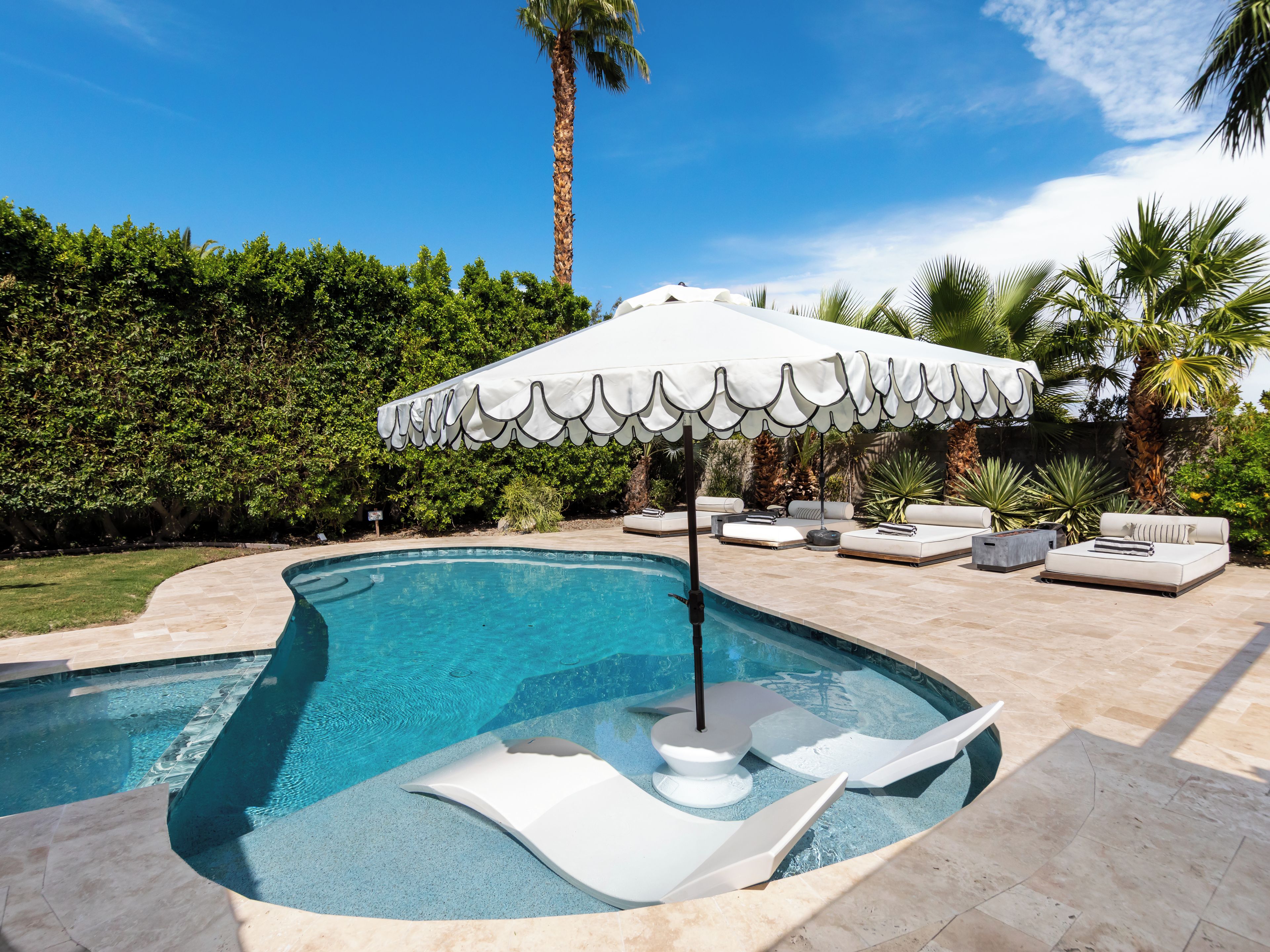 A swimming pool with a white umbrella, lounge chairs, and a palm tree in the background.