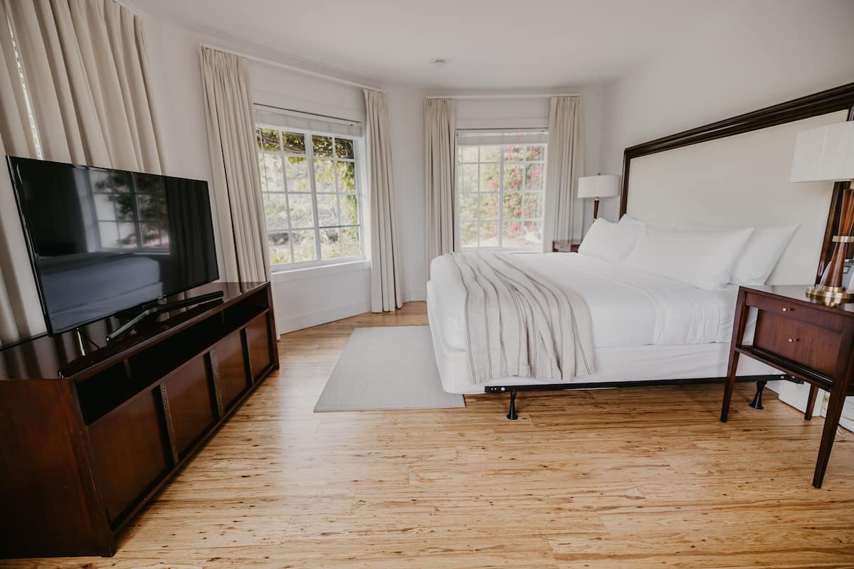 A bedroom with a large bed, a TV on a wooden cabinet, and windows with curtains.