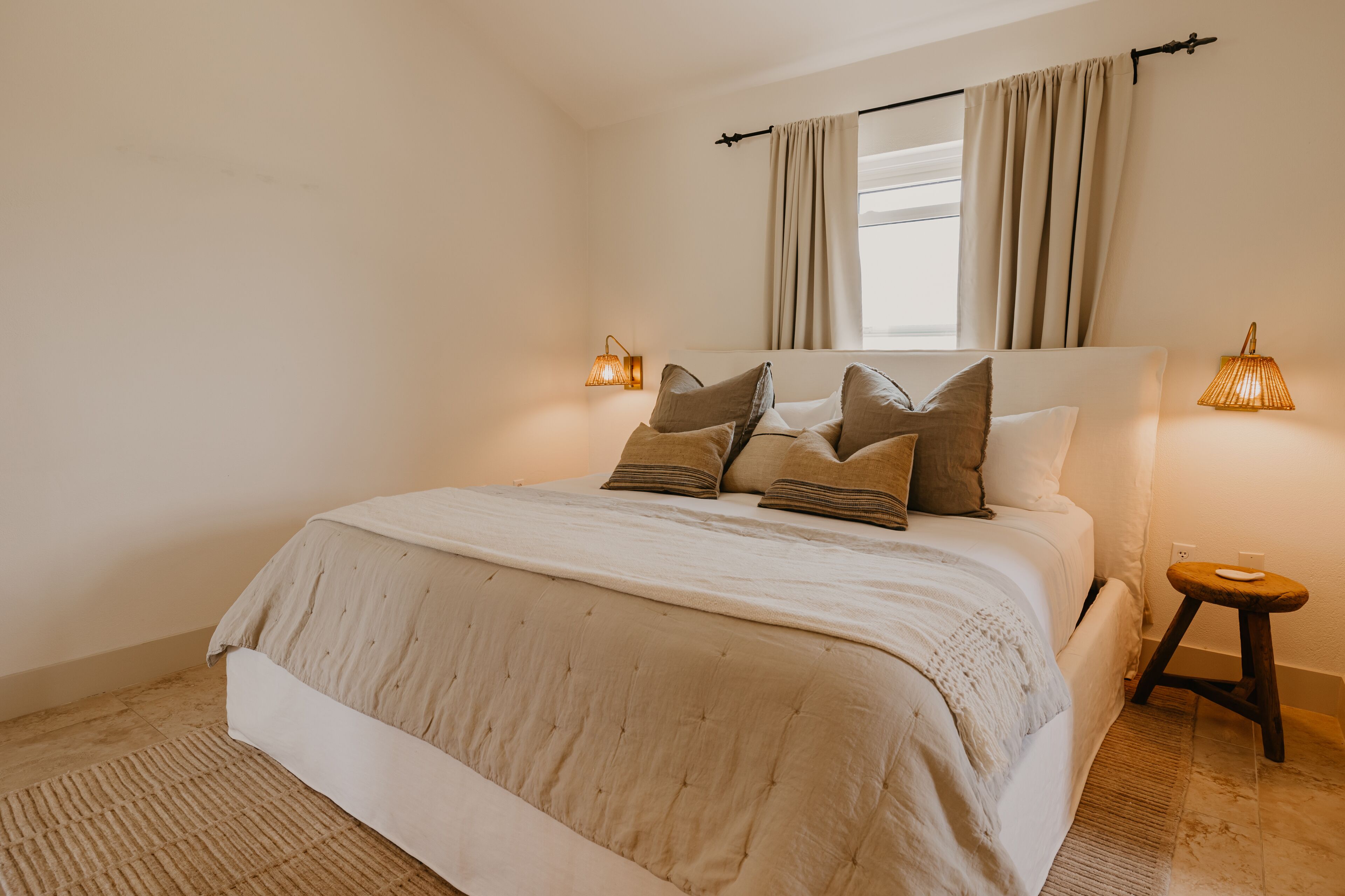 A bedroom with a large bed, a window with curtains, bedside lamps, and a small stool.