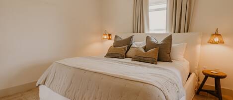 A bedroom with a large bed, a window with curtains, bedside lamps, and a small stool.