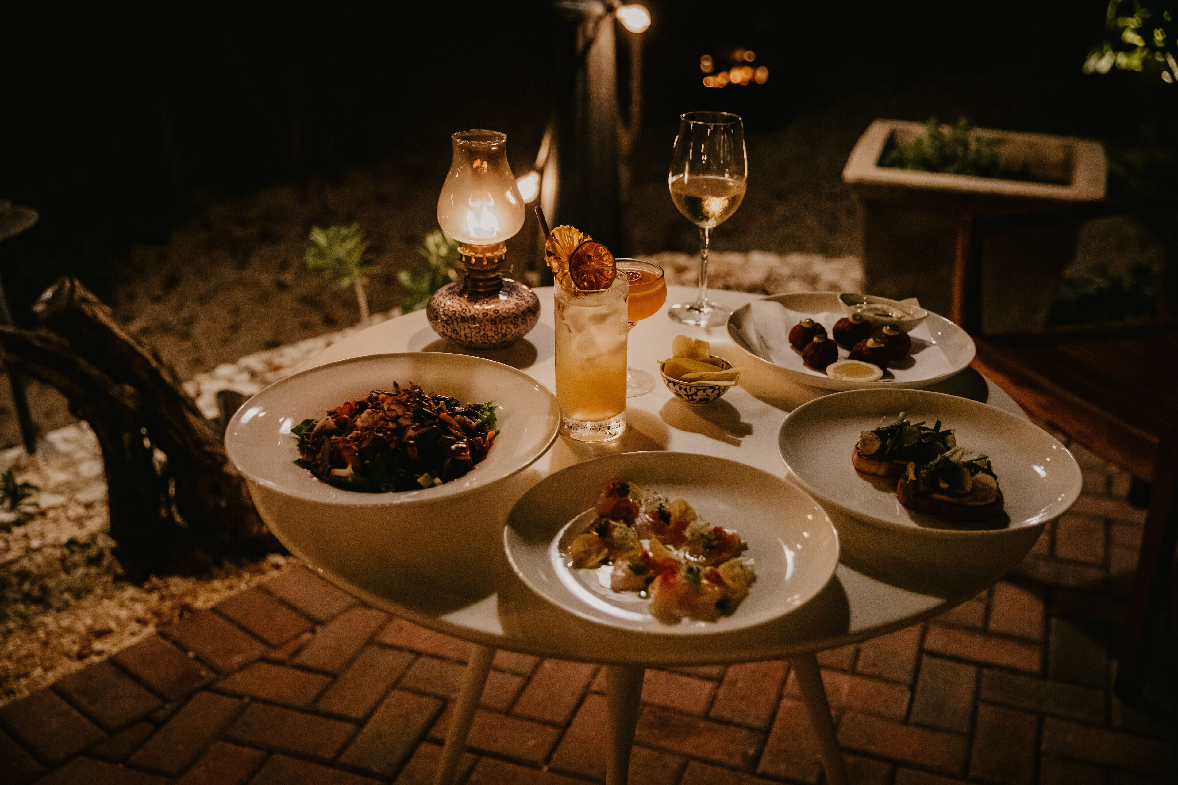 A table set outdoors with various dishes, a glass of wine, and a lit lamp.