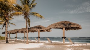 A beach with thatched umbrellas, lounge chairs, and palm trees. - Le Soleil d'Or CaymanBrac (West End)