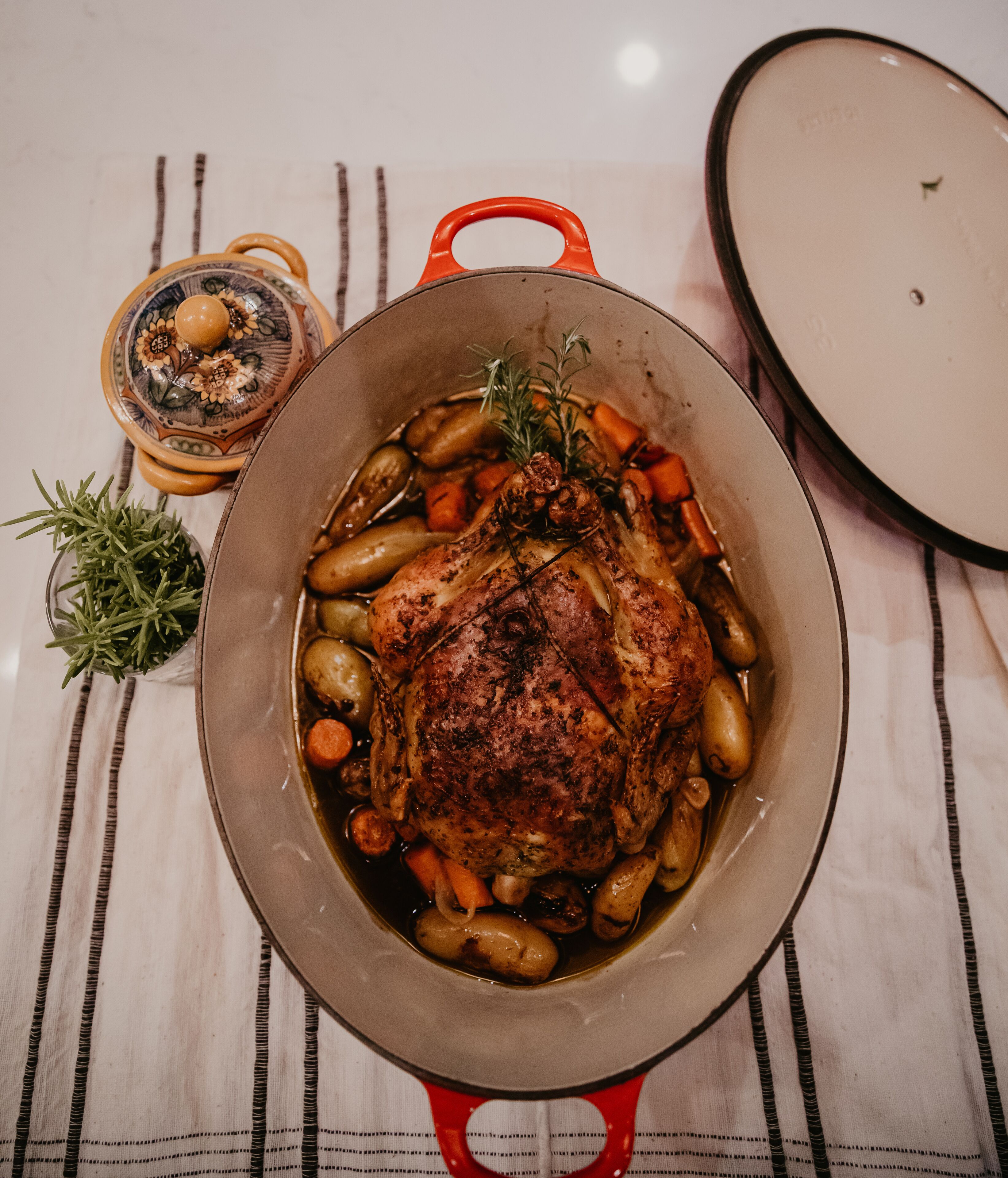 A roasted chicken in a pot with vegetables and herbs, placed on a striped tablecloth.