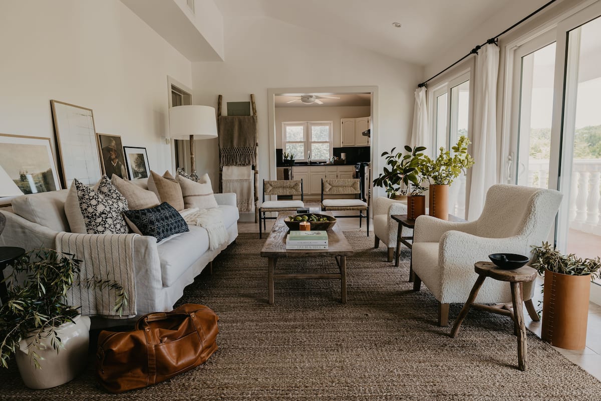 A spacious living room with a sofa, armchairs, a coffee table, and potted plants. A kitchen is visible in the background.