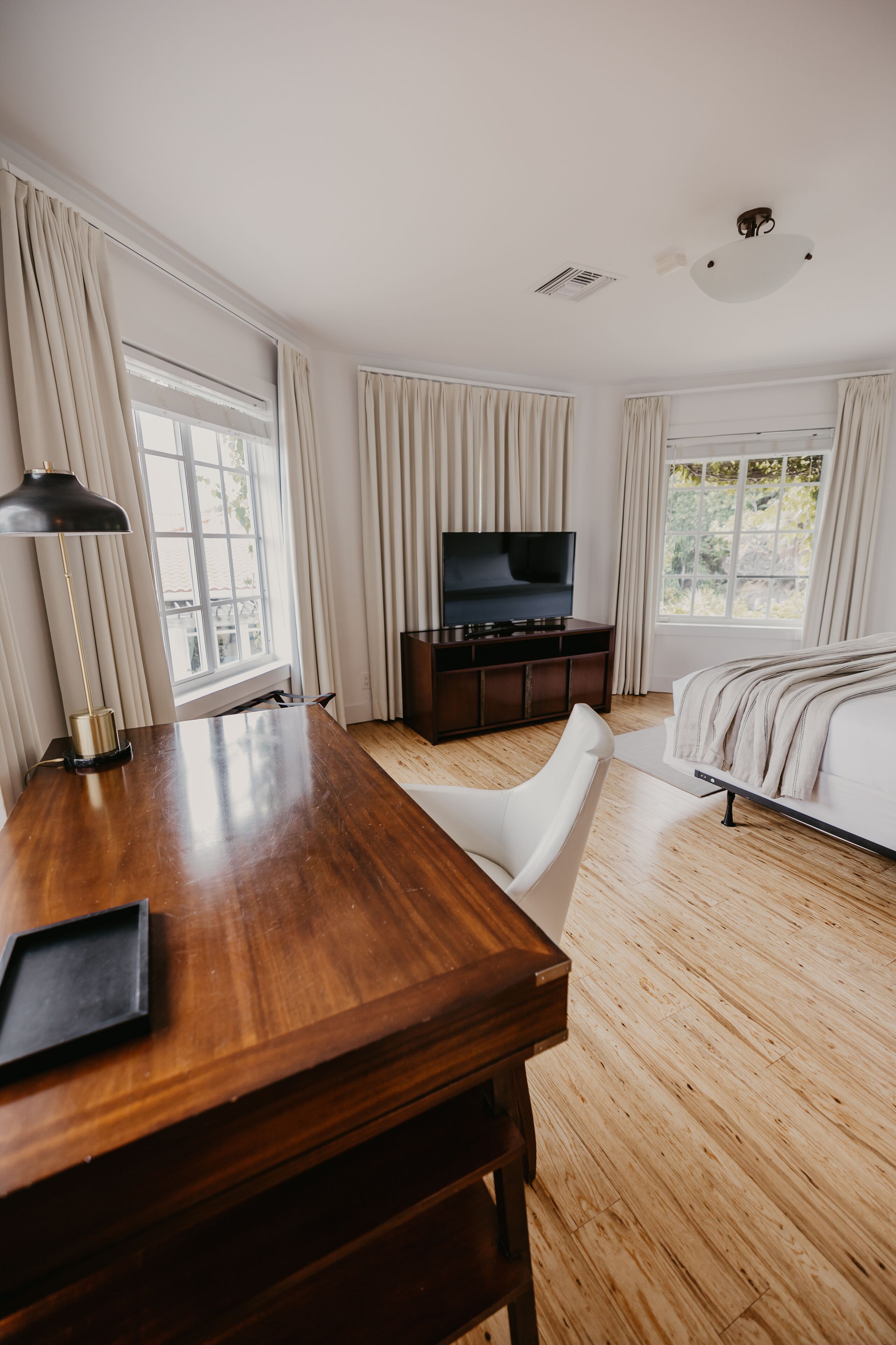 A hotel room with a wooden desk, a television on a cabinet, a bed with a white headboard, and large windows with curtains.