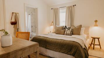 A bedroom with a bed, bedside lamps, a wooden table, a chair, a potted plant, and a window with curtains.