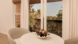 A round table with a bowl of fruit in a wicker chair, a window with white curtains, and a view of palm trees and a balcony. - Le Soleil d'Or CaymanBrac (West End)