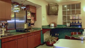 A kitchen with wooden cabinets, a refrigerator, a microwave, and a sink. There is a window with curtains and a potted plant on the counter. - Garden City Inn (Garden City)