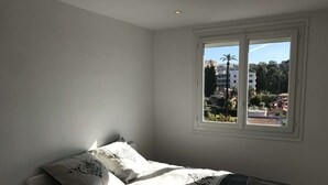 A bedroom with a bed, pillows, a window, and a view of buildings and trees. - Apartment 300 m from Cannes beach (Cannes)