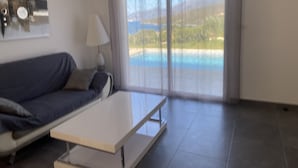 A modern living room with a sofa, a coffee table, and a view of a pool and landscape through large windows. - 2-standing seaside villa, breathtaking view of the Gulf of Saint Florent (Saint-Florent)