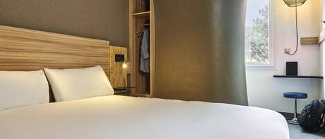 A hotel room with a large bed, a wooden headboard, a built-in wardrobe, a hanging light fixture, and a window with a view.