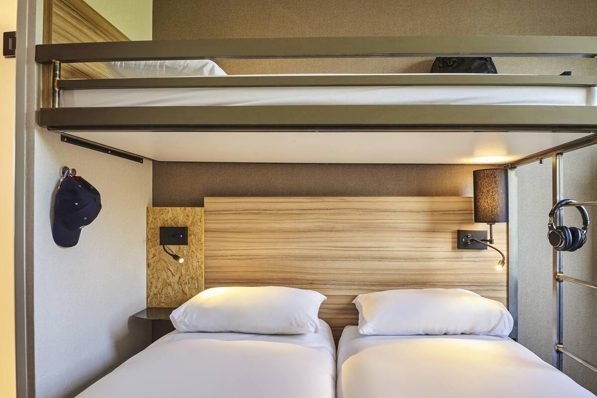 Een slaapkamer met een stapelbed, twee bedden, een houten hoofdbord, een wandlamp en een paar koptelefoons die aan de muur hangen.