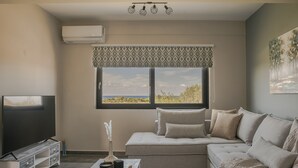 Ein modernes Wohnzimmer mit einem großen Flachbildfernseher, einem grauen Sofa mit vielen Kissen, einem Holztisch und einem Fenster mit Blick ins Freie.
