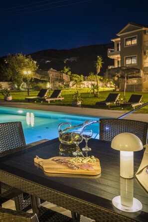 Un espace repas aménagé au bord de la piscine, avec une table dressée pour deux personnes, une planche en bois sur laquelle sont disposés des aliments et une lampe.