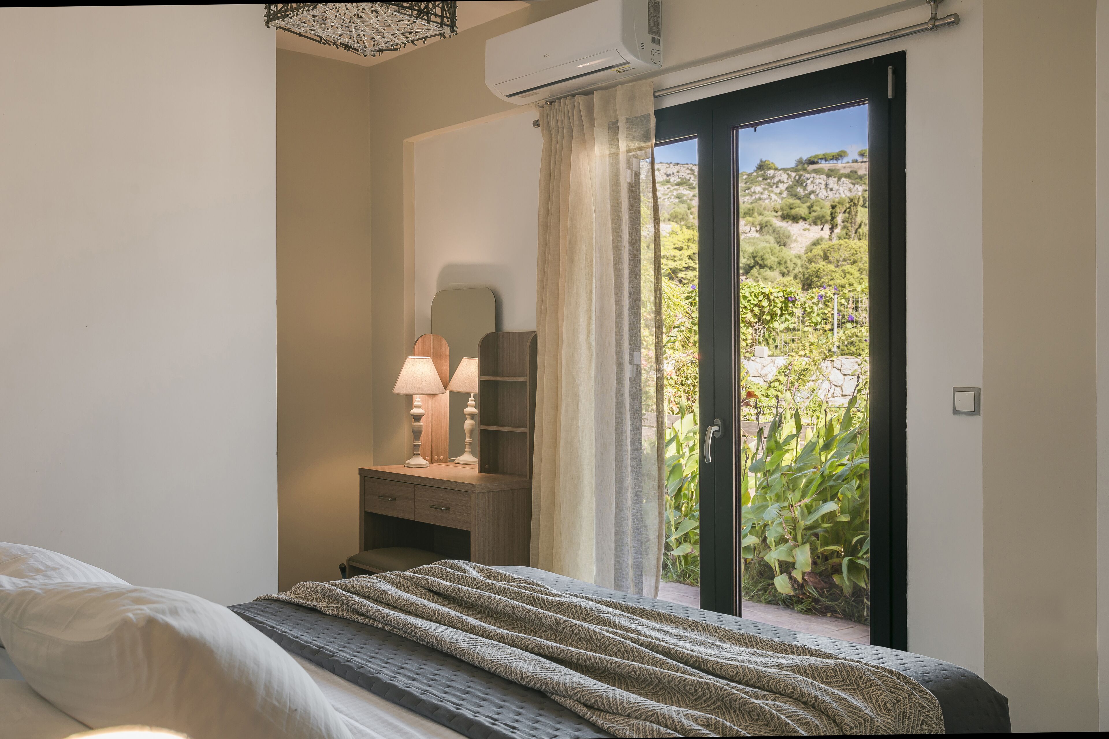 A bedroom with a bed, bedside table with lamps, a dresser, and a sliding glass door leading to a garden.