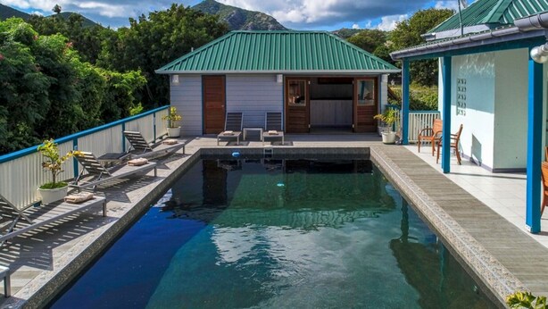 Ein Schwimmbecken mit Liegestühlen und einer überdachten Terrasse, umgeben von Grünpflanzen und Bergen.