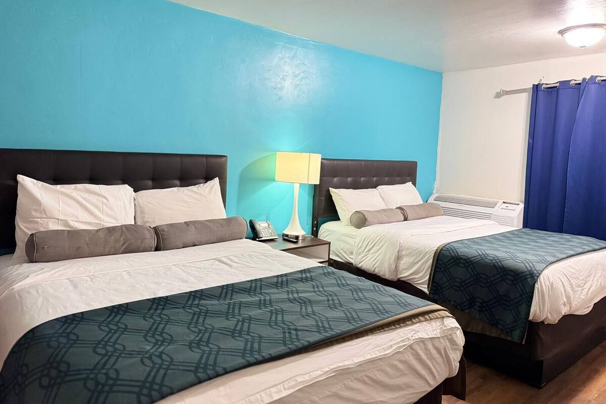 A hotel room with two beds, a nightstand with a lamp, and a window with blue curtains.