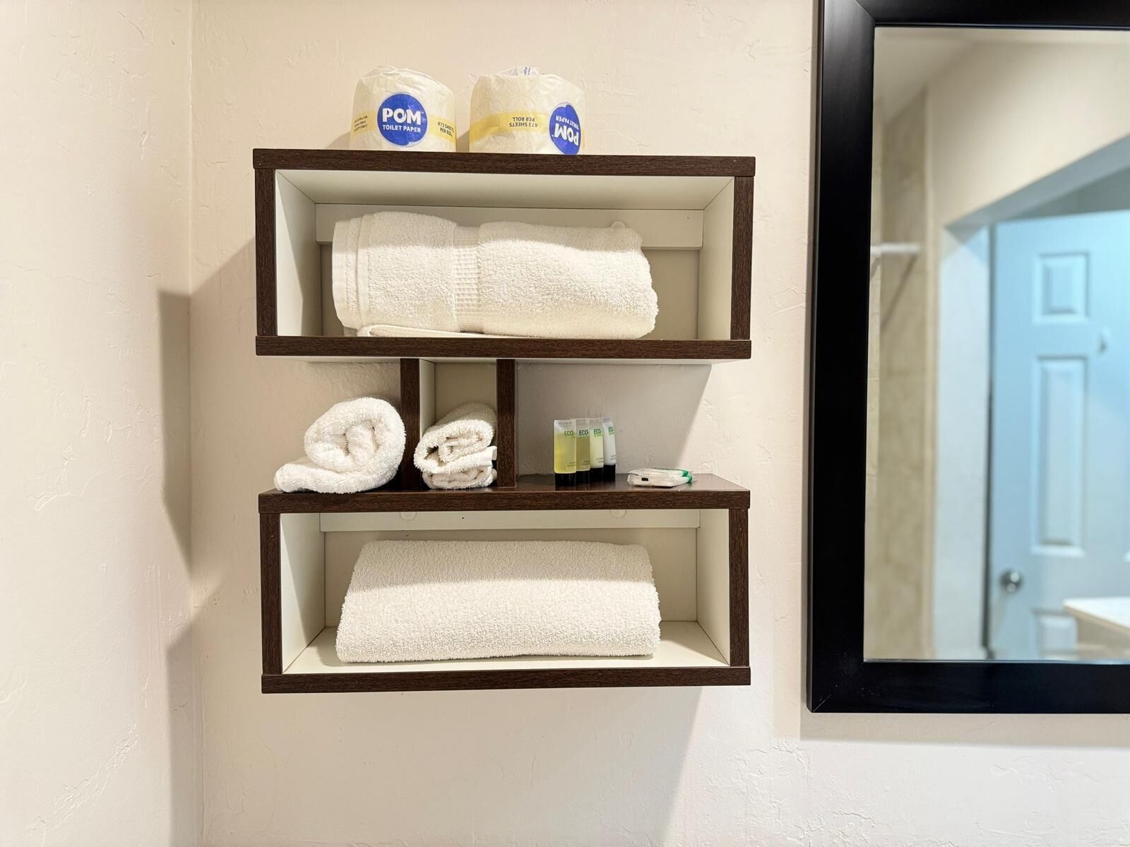 A bathroom with a shelf holding white towels, a bottle of POM toilet paper, and a bottle of hand soap.