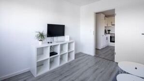 A modern, minimalist living room with a flat-screen TV, a small plant, and a laptop on a white entertainment unit. A grey wooden floor and a small white table are visible. - Vacation apartment Ankerplatz in House Annika in Schillig-Horumersiel (Wangerland)