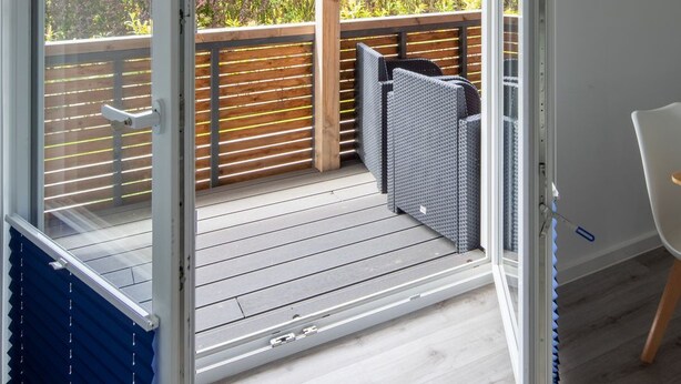 An open sliding door leading to a balcony with a wooden deck, a seating area with a chair, and a view of a garden. - Vacation apartment Ankerplatz in House Annika in Schillig-Horumersiel (Wangerland)