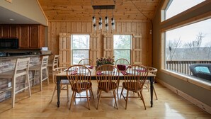 Un comedor de madera con una mesa grande y sillas, una lámpara colgante y ventanas amplias.