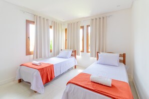 A room with two single beds, each with a white bedspread and an orange blanket, and white pillows. There are curtains on the windows, and the room has a tiled floor.