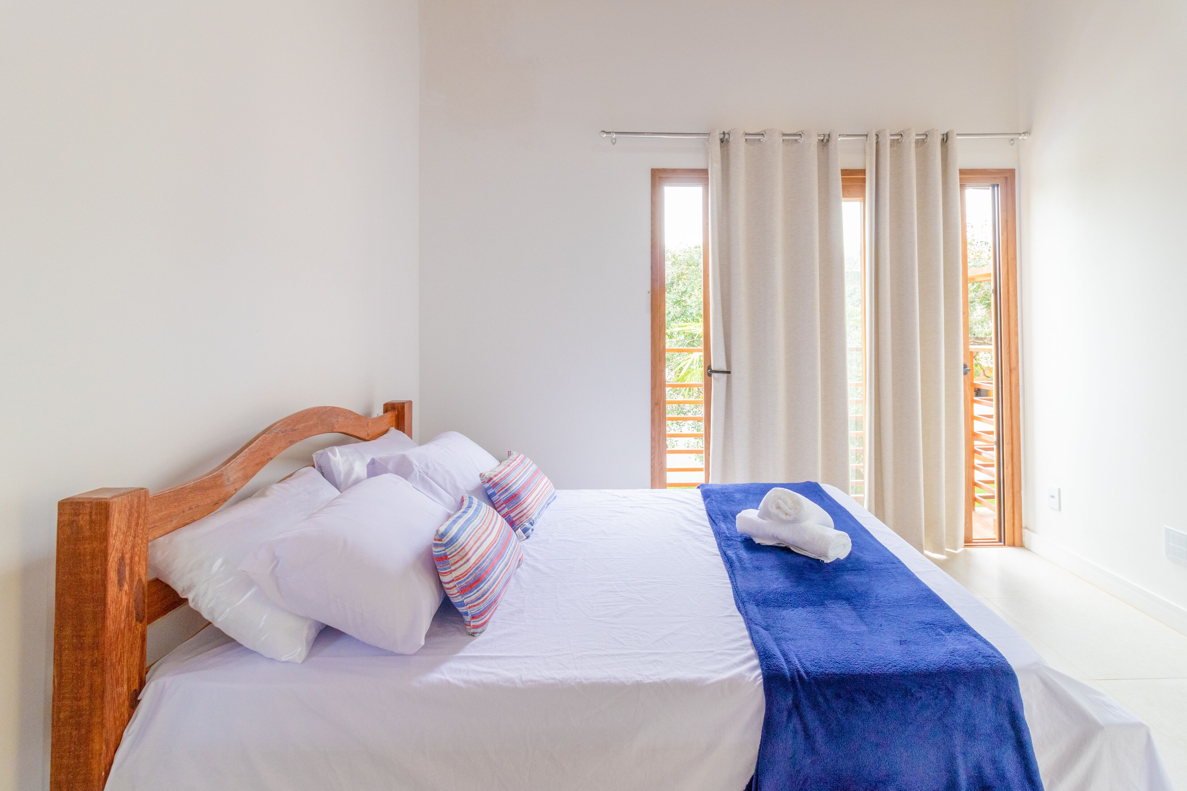 Ein ordentlich bezogenes Bett mit weißen Leinentüchern und einer blauen Decke, ein Holz-Kopfteil und ein Fenster mit Vorhängen.