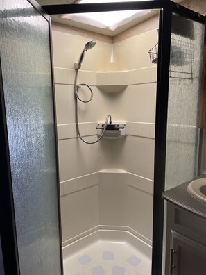 Une douche d’angle avec une porte en verre, une douchette à main et une petite étagère avec un crochet.