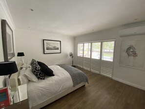 A bedroom with a bed, a bedside table with a lamp, a framed picture on the wall, and a window with blinds.