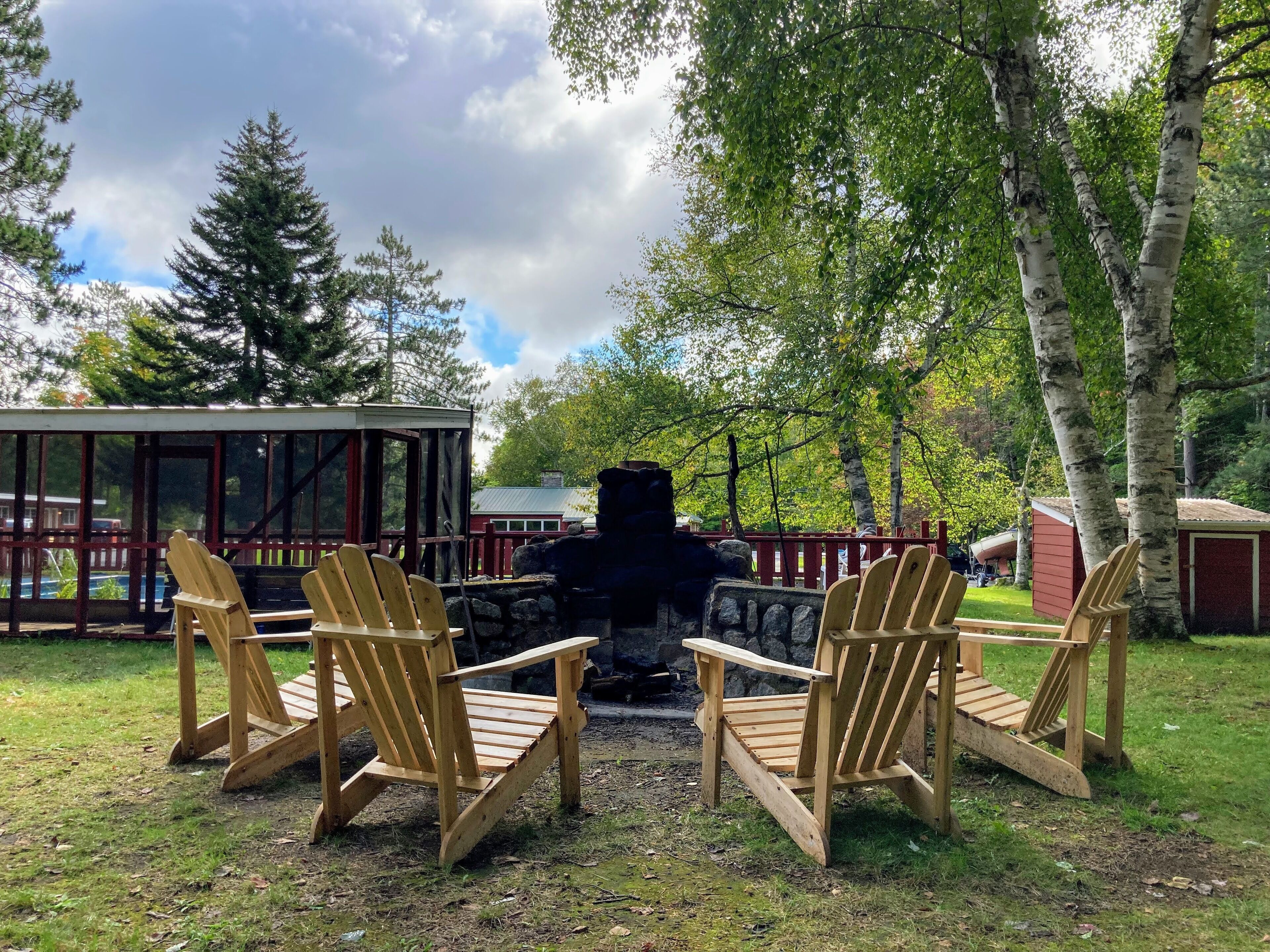 Une terrasse en bois avec des fauteuils Adirondack donnant sur une cheminée en pierre, le tout entouré d’arbres et d’une structure en verre.
