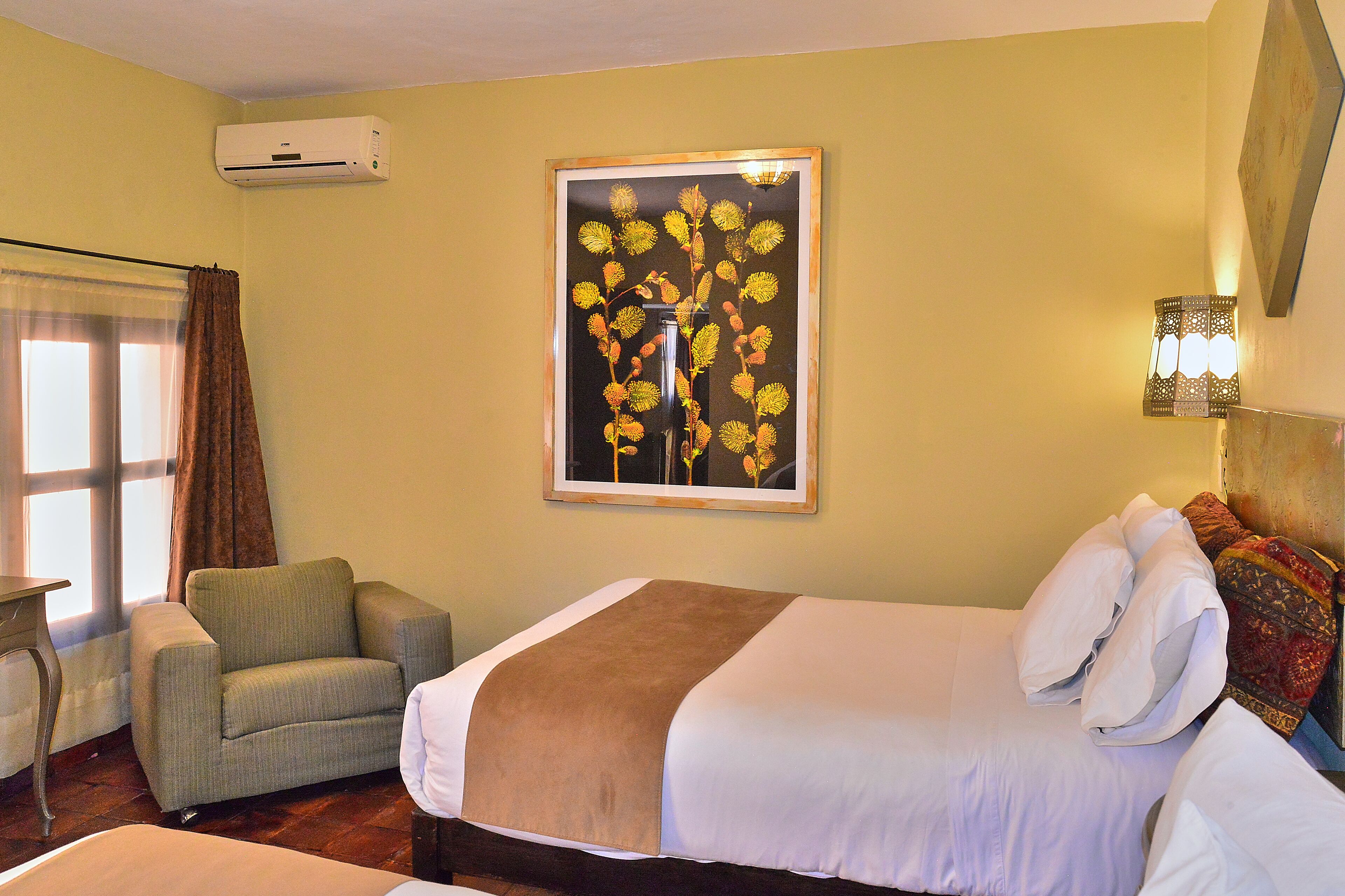 A hotel room with a bed, a chair, a wall-mounted air conditioner, and a framed picture of a plant.
