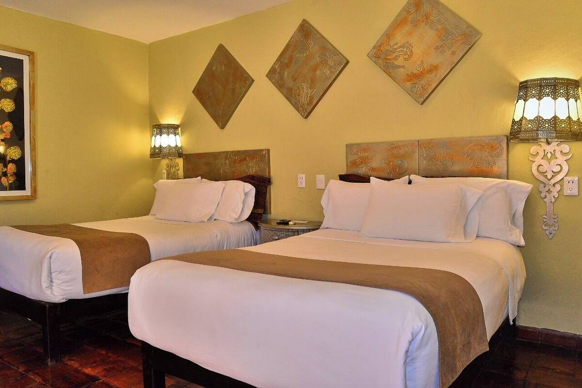 A hotel room with two beds, a painting, and wall-mounted lamps.