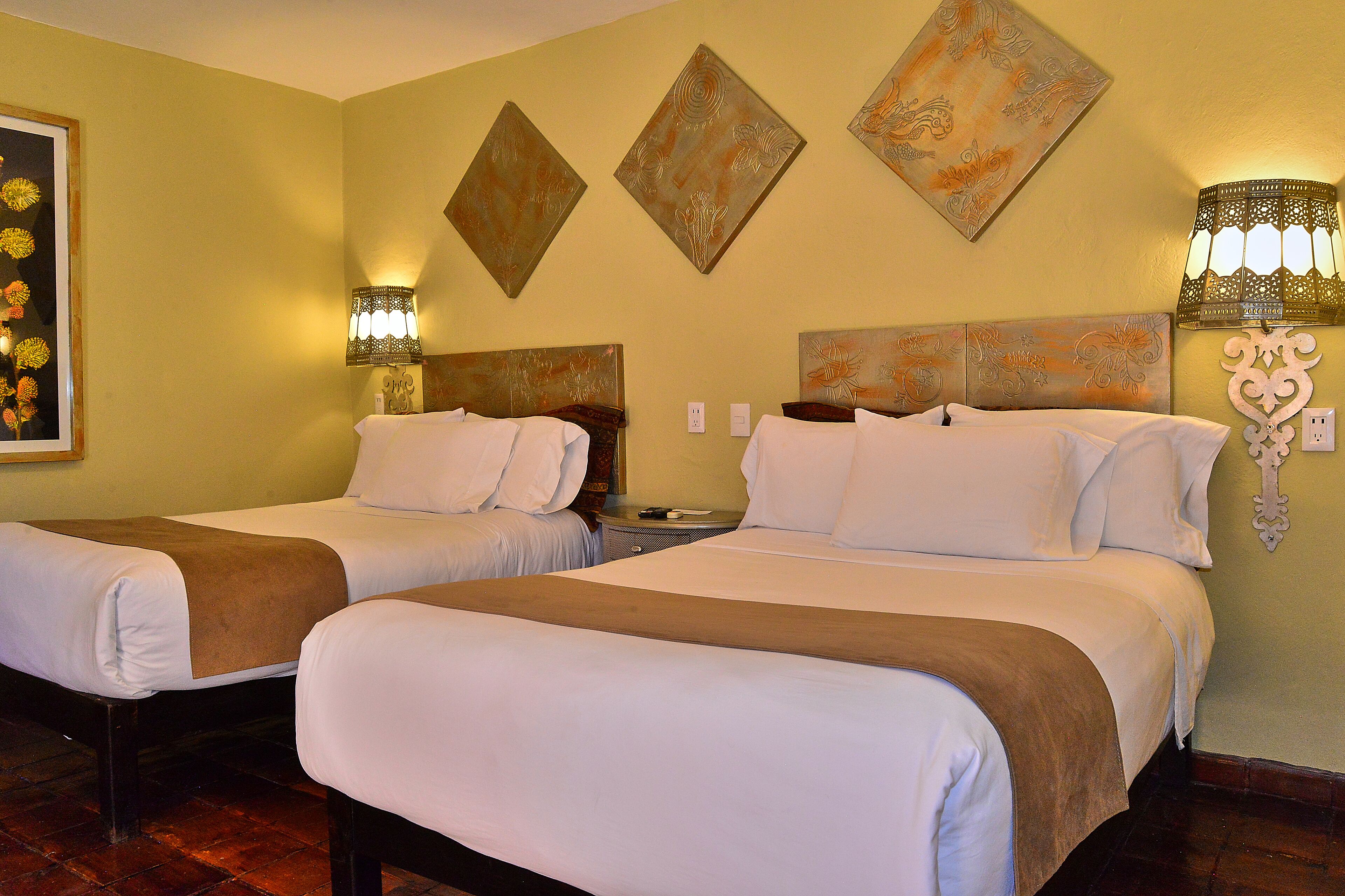 Habitación de hotel con dos camas, cada una con ropa de cama blanca y cabeceras marrones. Hay lámparas de pared a cada lado de las camas, y un cuadro de flores en la pared izquierda.