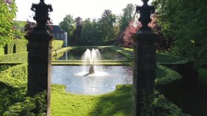 Ein friedlicher Garten mit einem Brunnen, ordentlich geschnittenen Hecken und einem Gebäude in der Ferne.