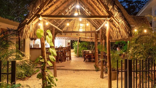 A thatched-roof structure with a gravel pathway leading to a dining area, surrounded by greenery. - Palm Beach Inn (Cabo Velas)